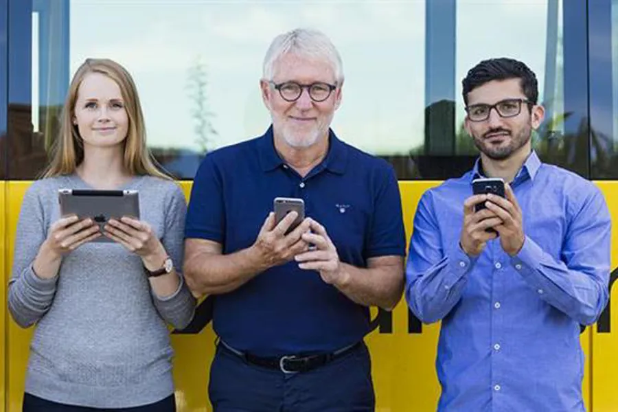 Tre personer med mobiler og nettbrett utenfor en buss. Foto.