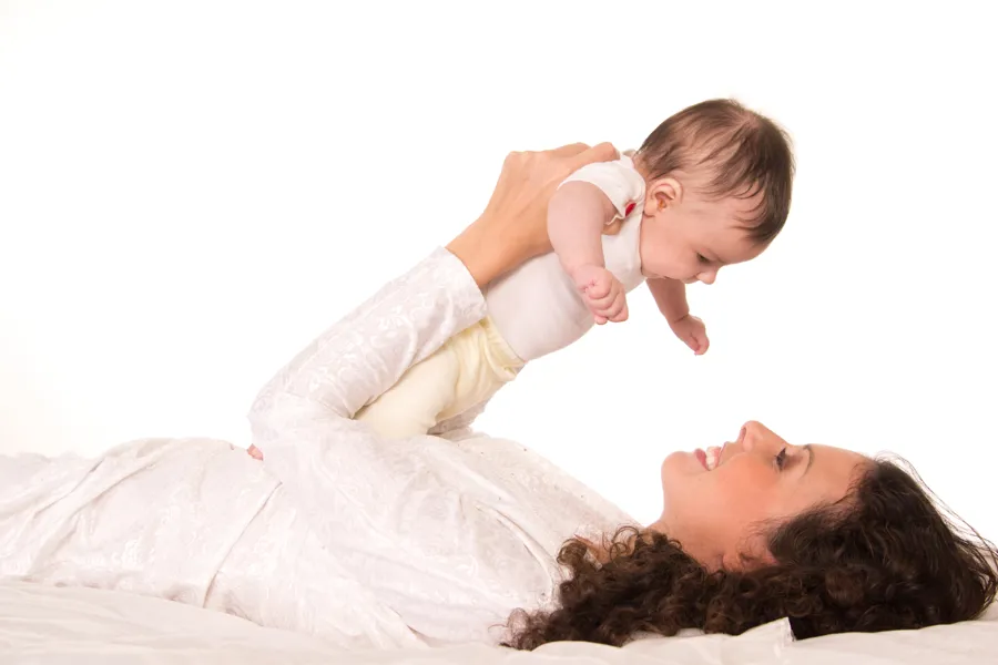 Kvinne ligger i en seng og holder barnet sitt over sitt eget ansikt. Foto